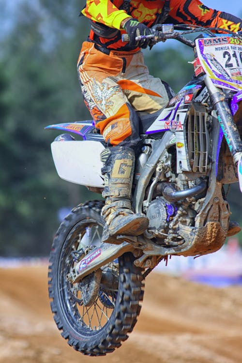 Person in Orange and Yellow Fox Motorcycle Suit Riding a Purple White Gray and Black Dirt Motorcycle Outdoors during Daytime