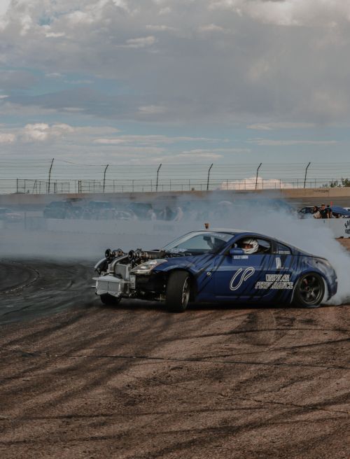 Free Dynamic shot of a blue Nissan 350Z performing a drift on a dusty racetrack with smoke trailing. Stock Photo