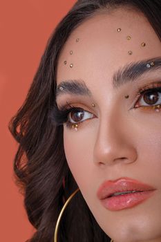 Close-up portrait of a woman with gemstone makeup on orange background, showcasing elegant beauty and creativity.