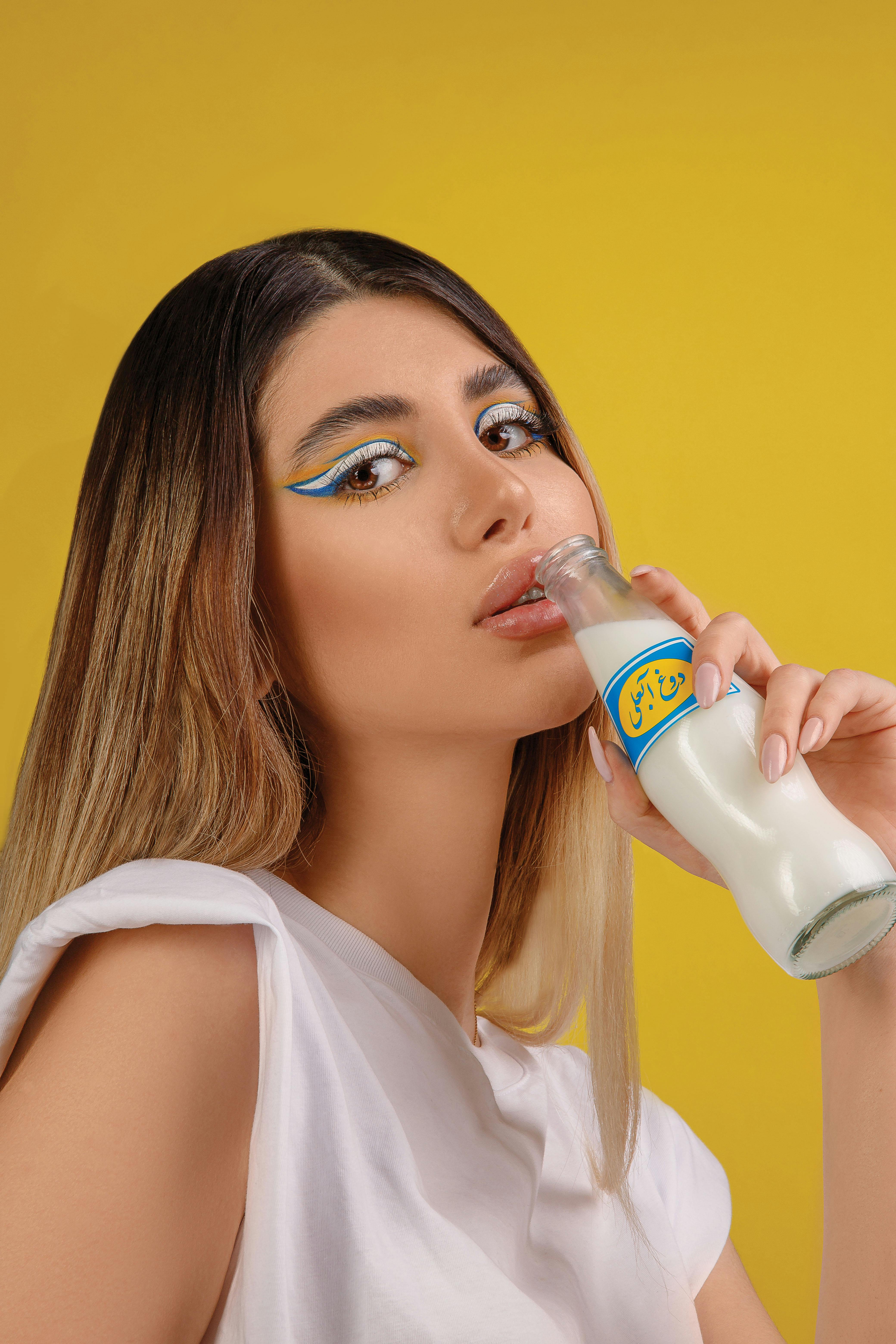 Chic woman with bold eye makeup holding a milk bottle against a vibrant yellow backdrop.