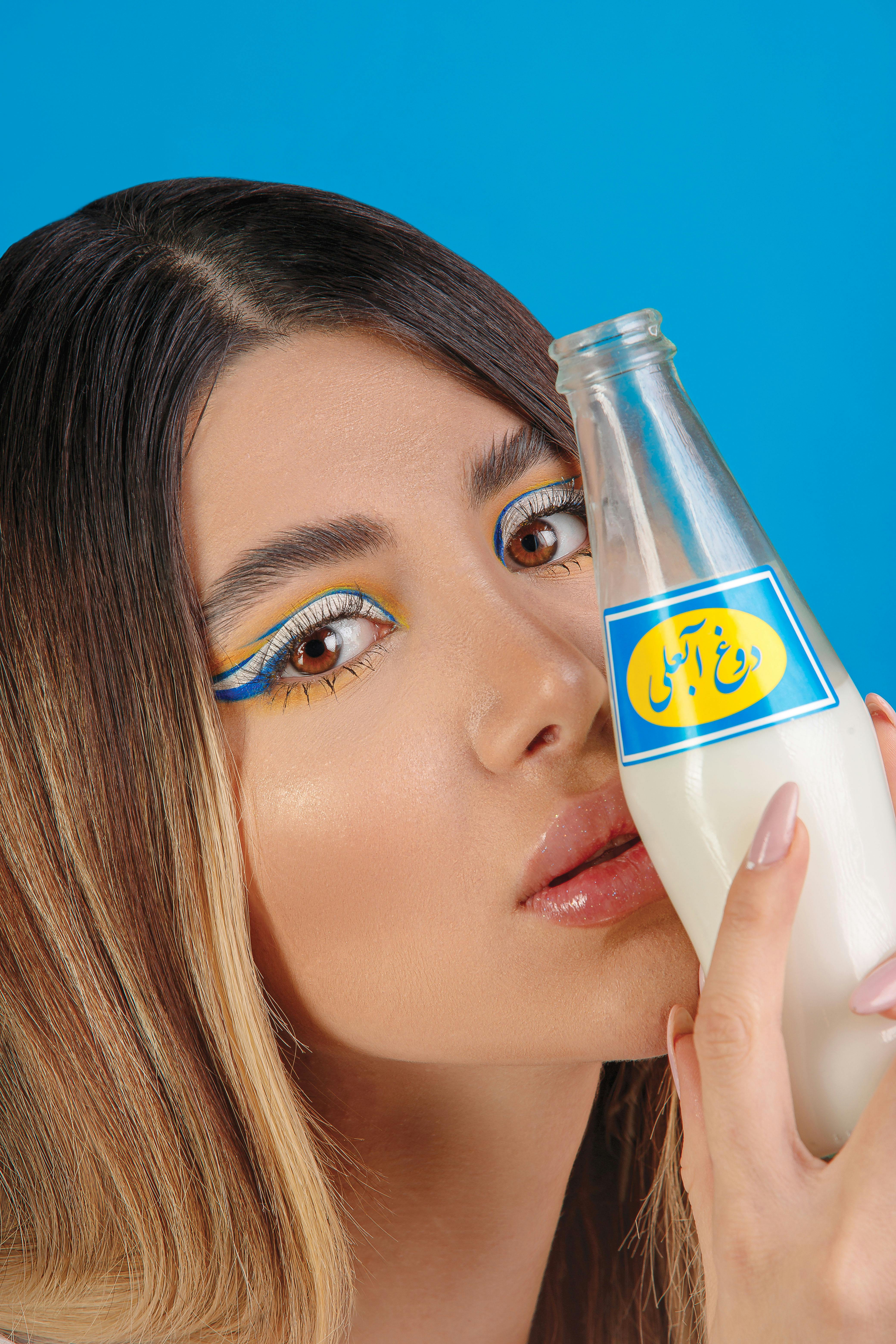 Modern portrait of a woman with artistic makeup holding a milk bottle against a blue background.