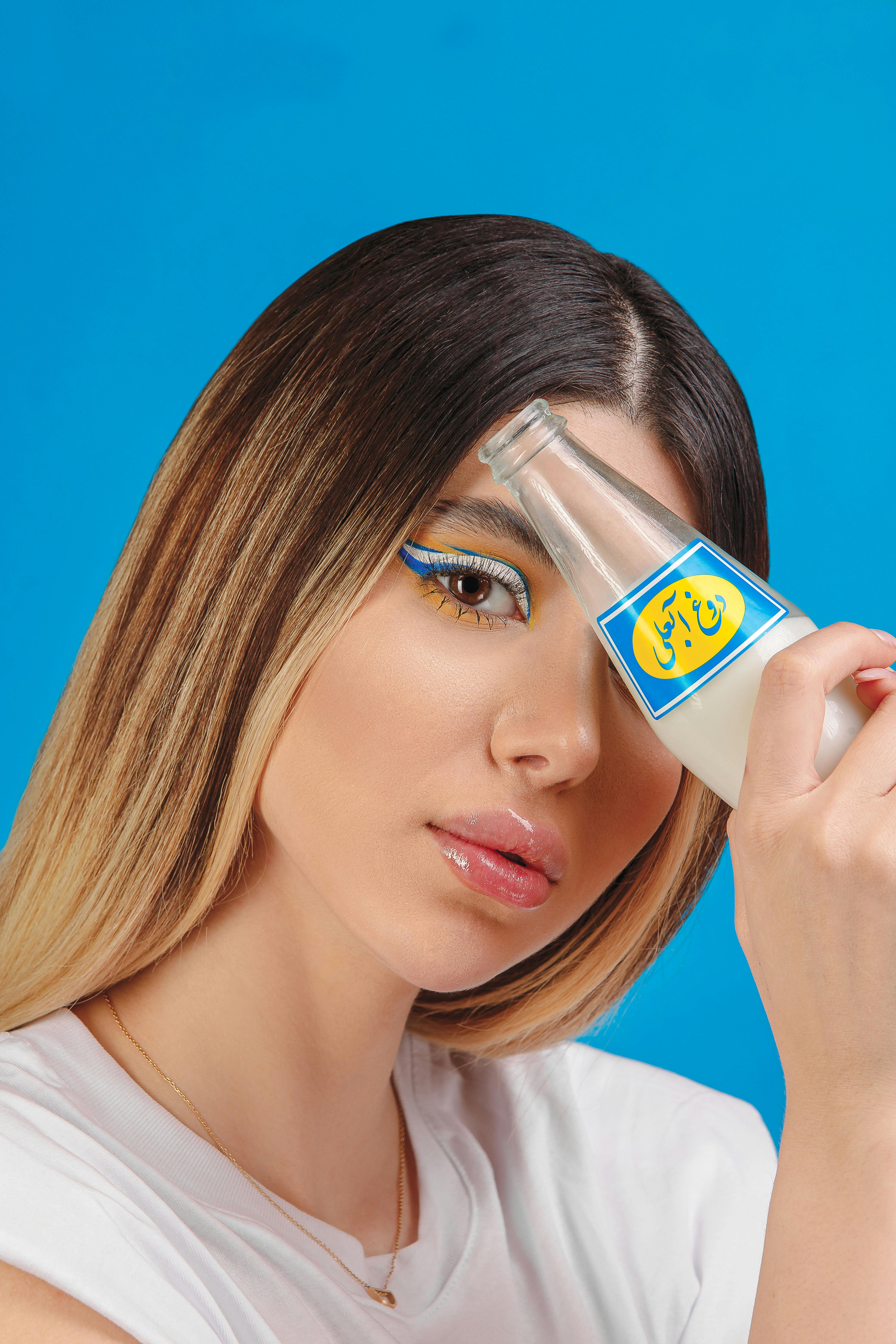 Stylish woman with unique makeup holding a glass bottle against a blue backdrop.