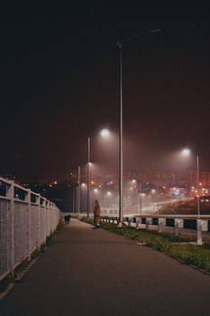 A lone pedestrian walks along a foggy city sidewalk under dim streetlights at night.