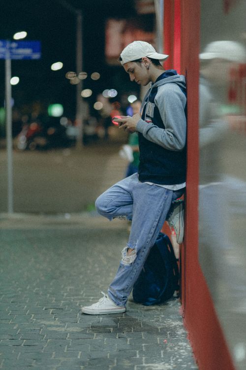 Free Young man in casual streetwear using a smartphone at night outdoors, leaning against a wall. Stock Photo