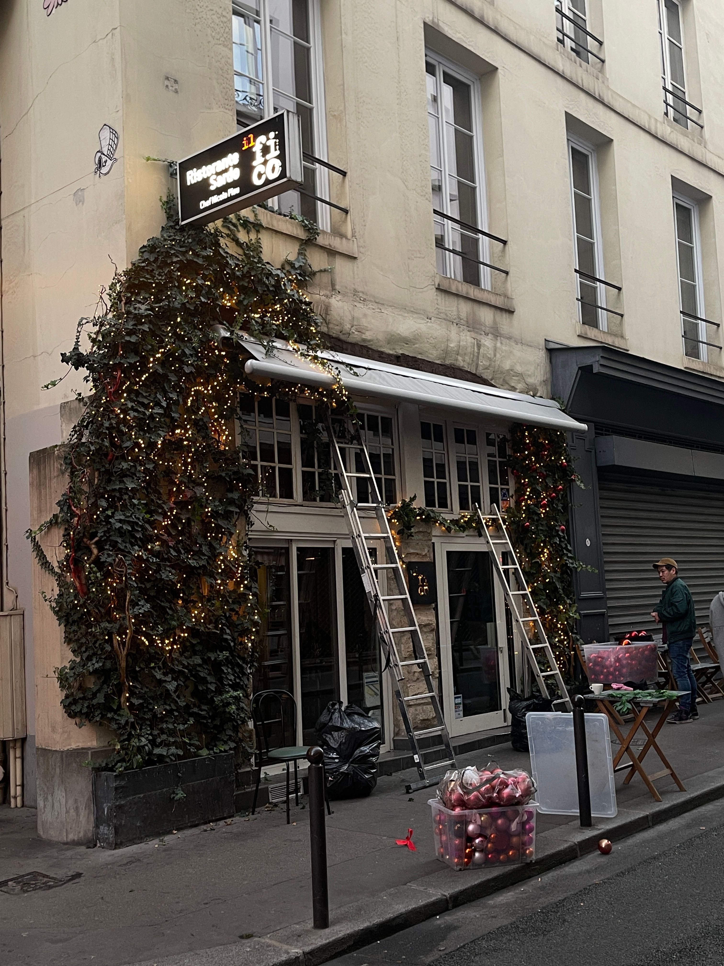 Festive Christmas decorations on a restaurant facade in an urban street setting, featuring dangling lights and greenery.
