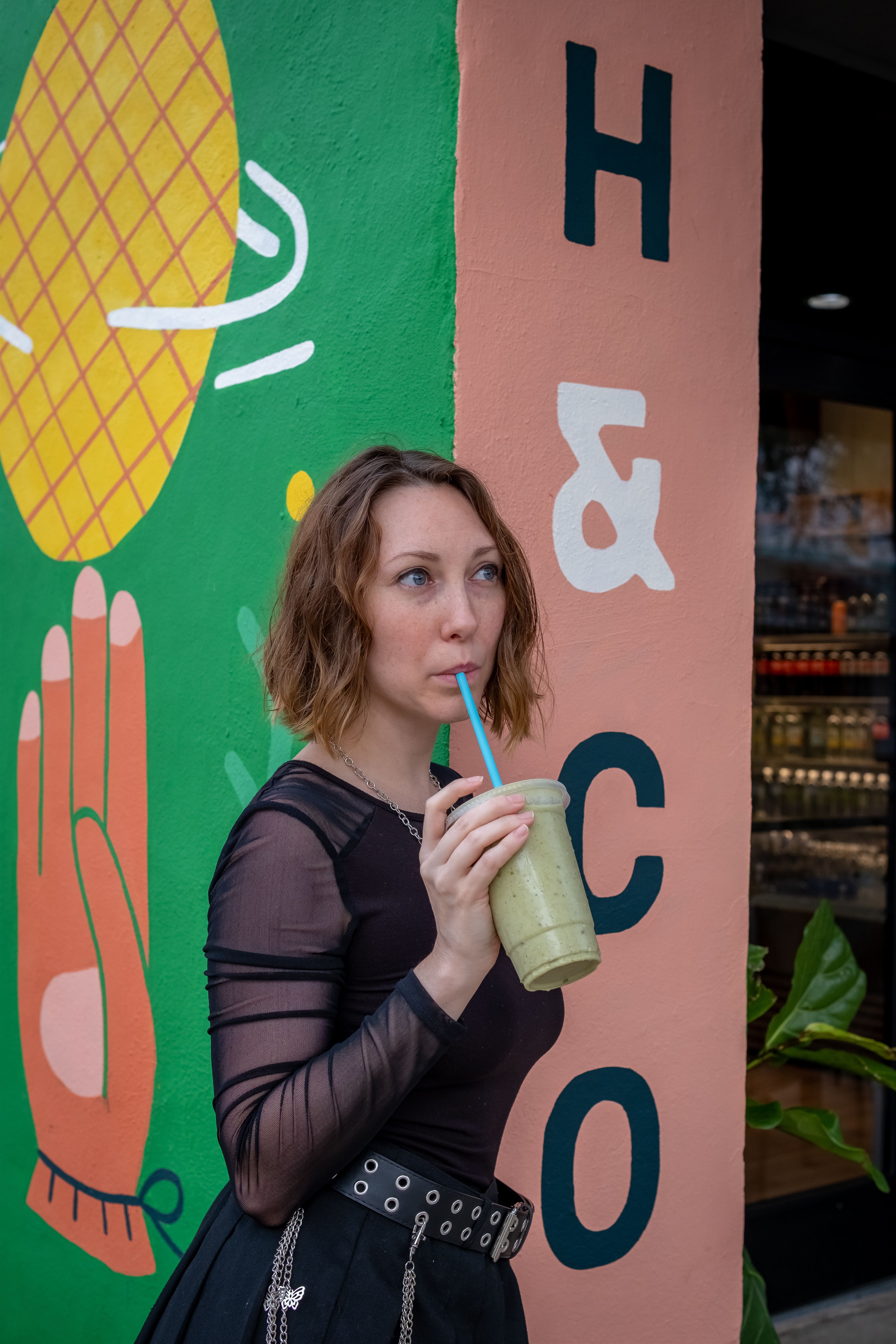 Stylish woman sipping a green smoothie in front of a colorful mural on a sunny day.