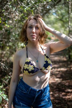 A young woman in a floral bikini top and denim shorts poses in a sunlit forest path.