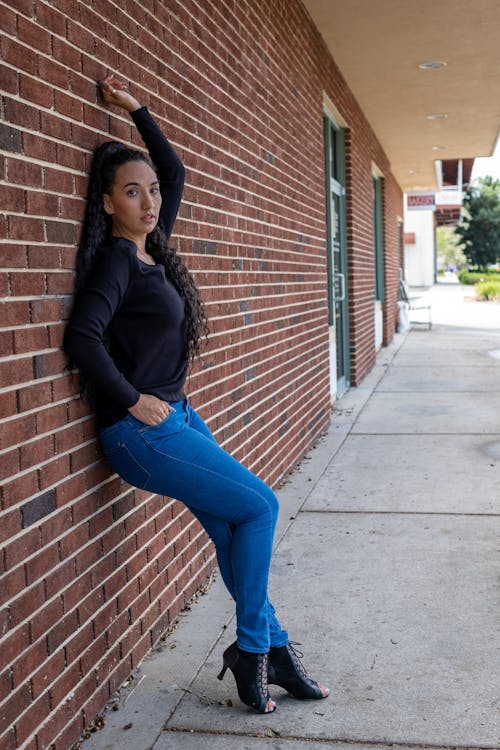 A Woman in a Black Long Sleeves and Denim Pants Leaning on a Brick Wall