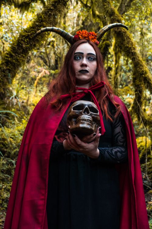 Free A woman in a red cape holds a skull amidst a spooky forest, embodying mystery and fantasy. Stock Photo
