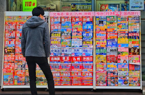 Free Standing Man Wearing Gray Hoodie in Front of Magazine in Rack Stock Photo