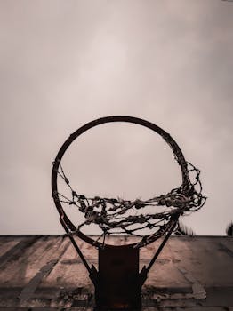 Low-angle view of an abandoned basketball hoop with tattered netting set against an overcast sky.