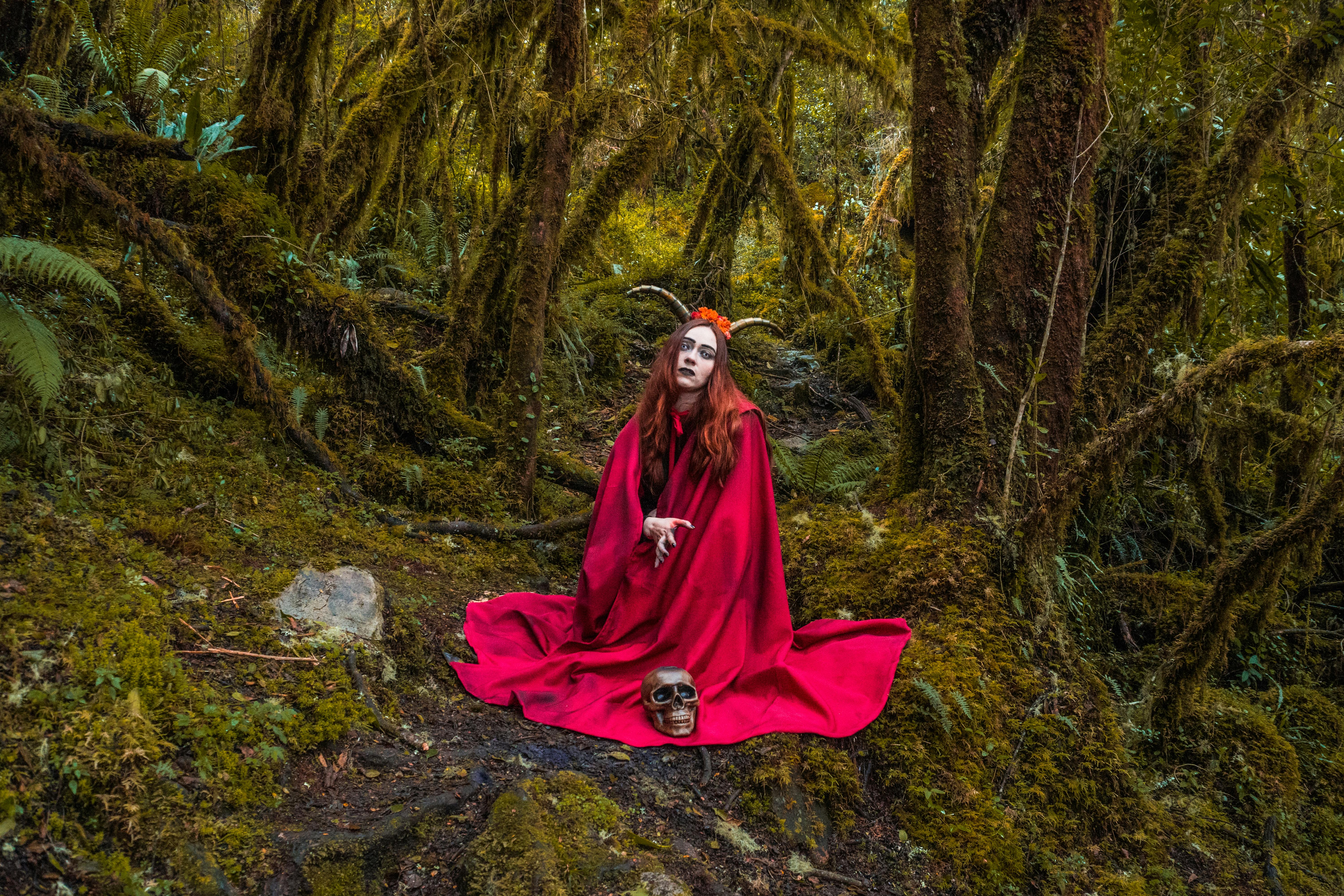A mysterious witch in a red gown sits in a forest with a skull, evoking fantasy and mystery.