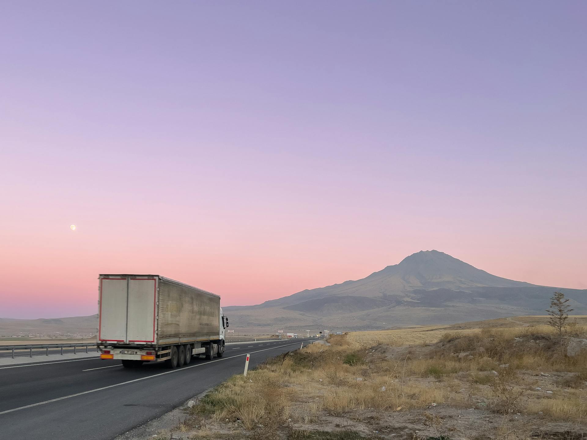 A lone truck drives on a highway against a beautiful pink dusk sky with a mountain in the background.