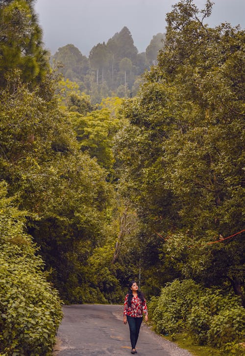 A Woman Walking on the Road Between Green Trees