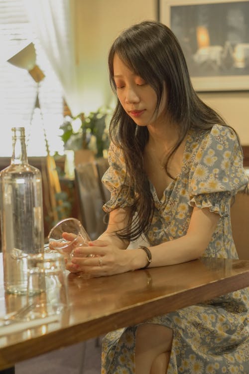 A Woman in Floral Dress Sitting Near the Wooden Table while Holding a Drinking Glass