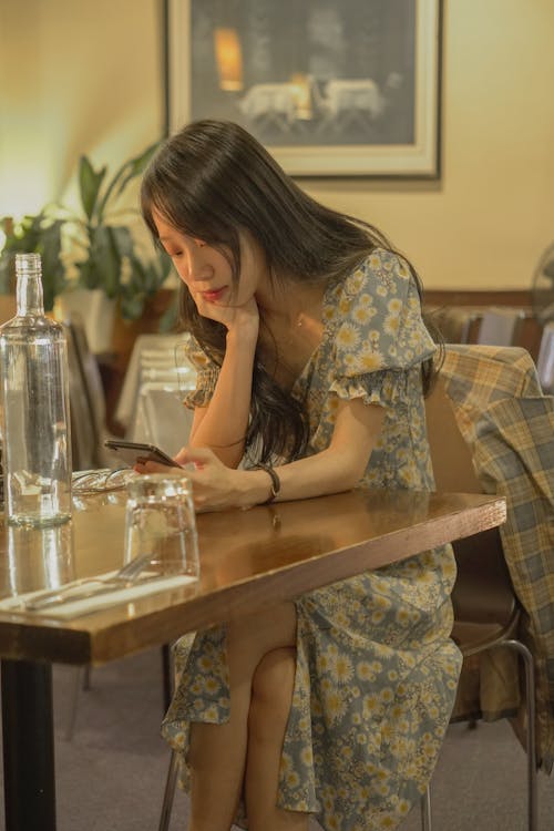 A Woman in Floral Dress Sitting Near the Wooden Table while Using Her Phone