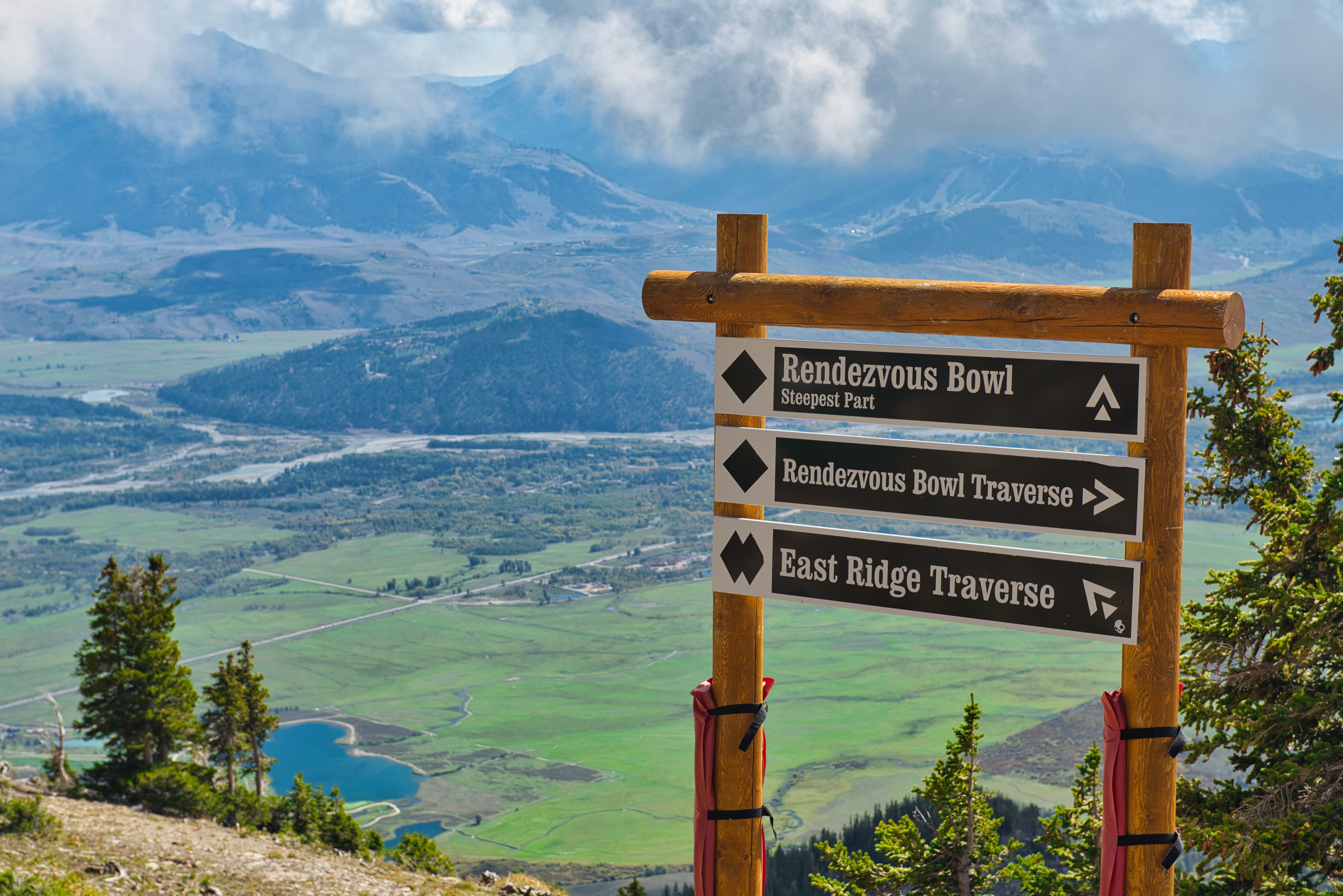 Panoramic view of Jackson, WY with a wooden signpost pointing towards Rendezvous Bowl and East Ridge Traverse.