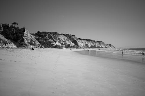 A Grayscale Photo of People Walking on the Beach Near the Rock Formation