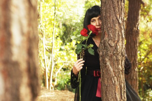 Mysterious woman in an elegant dress holding red roses, partially hidden behind a tree in a bright forest.