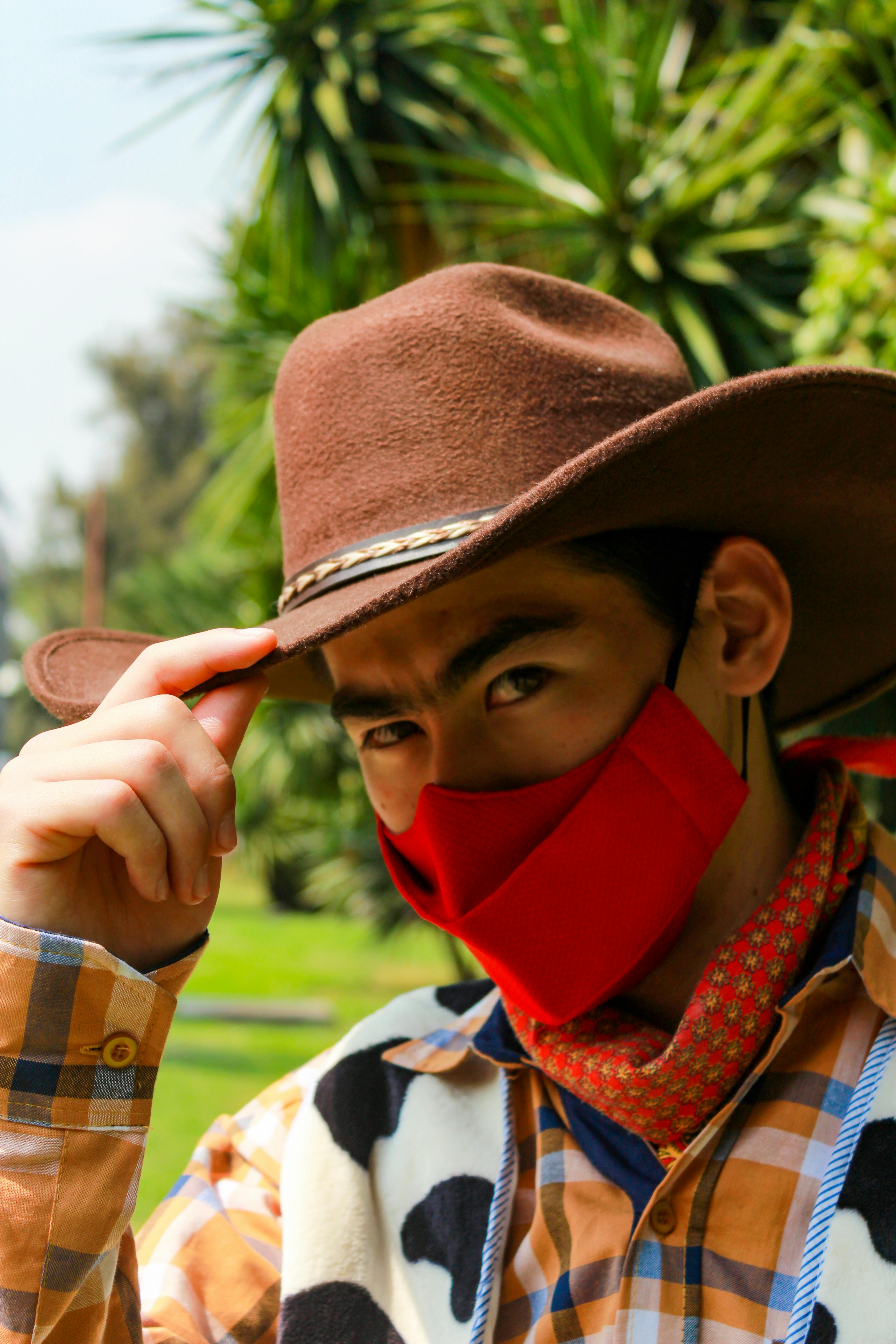 Portrait of a young man in cowboy attire wearing a red face mask outdoors, representing new normal.