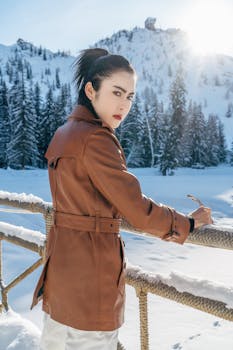 Woman in a brown coat on a snowy bridge with mountains in the background.