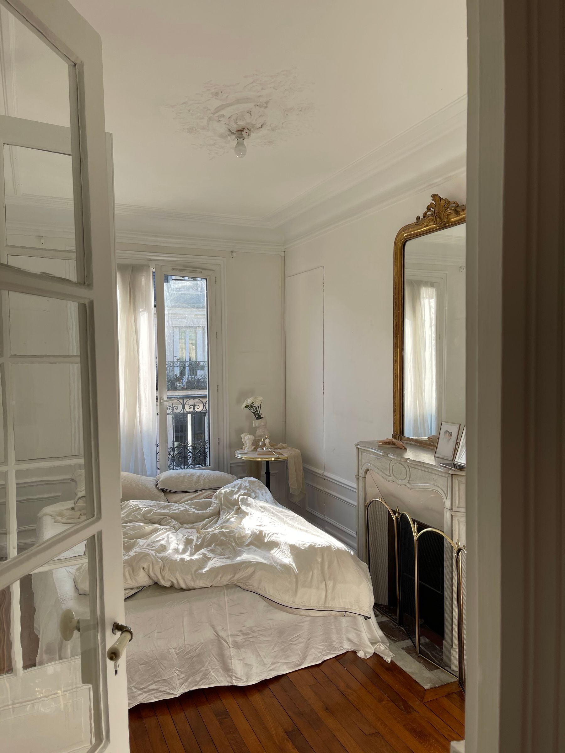 French-style bedroom with white linen bed, carved fireplace, gilded mirror, timber floor and glazed balcony doors