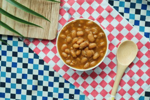 An overhead shot of baked beans in a bowl with a wooden spoon on a colorful checkered tablecloth.