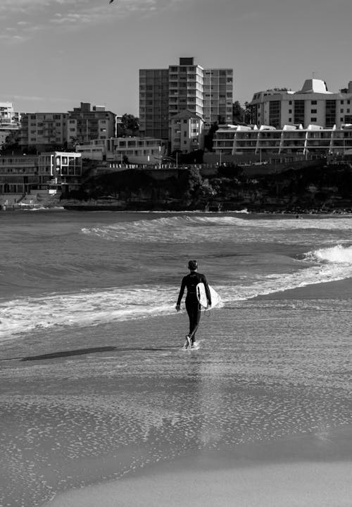 Black and White Photo of Man Walking on Shore While Carrying a Surfboard