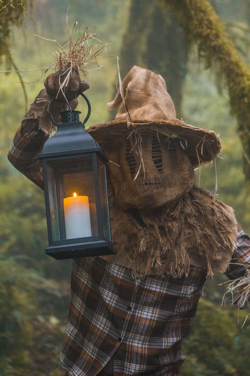 Person Dressed in a Scarecrow Halloween Costume Holding a Lantern in a Forest 