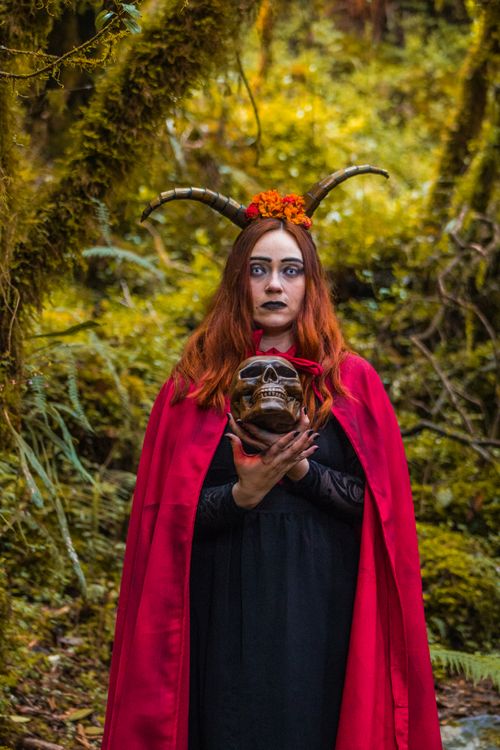 Free A woman in a red cape and horns holds a skull in a mystical forest setting. Stock Photo