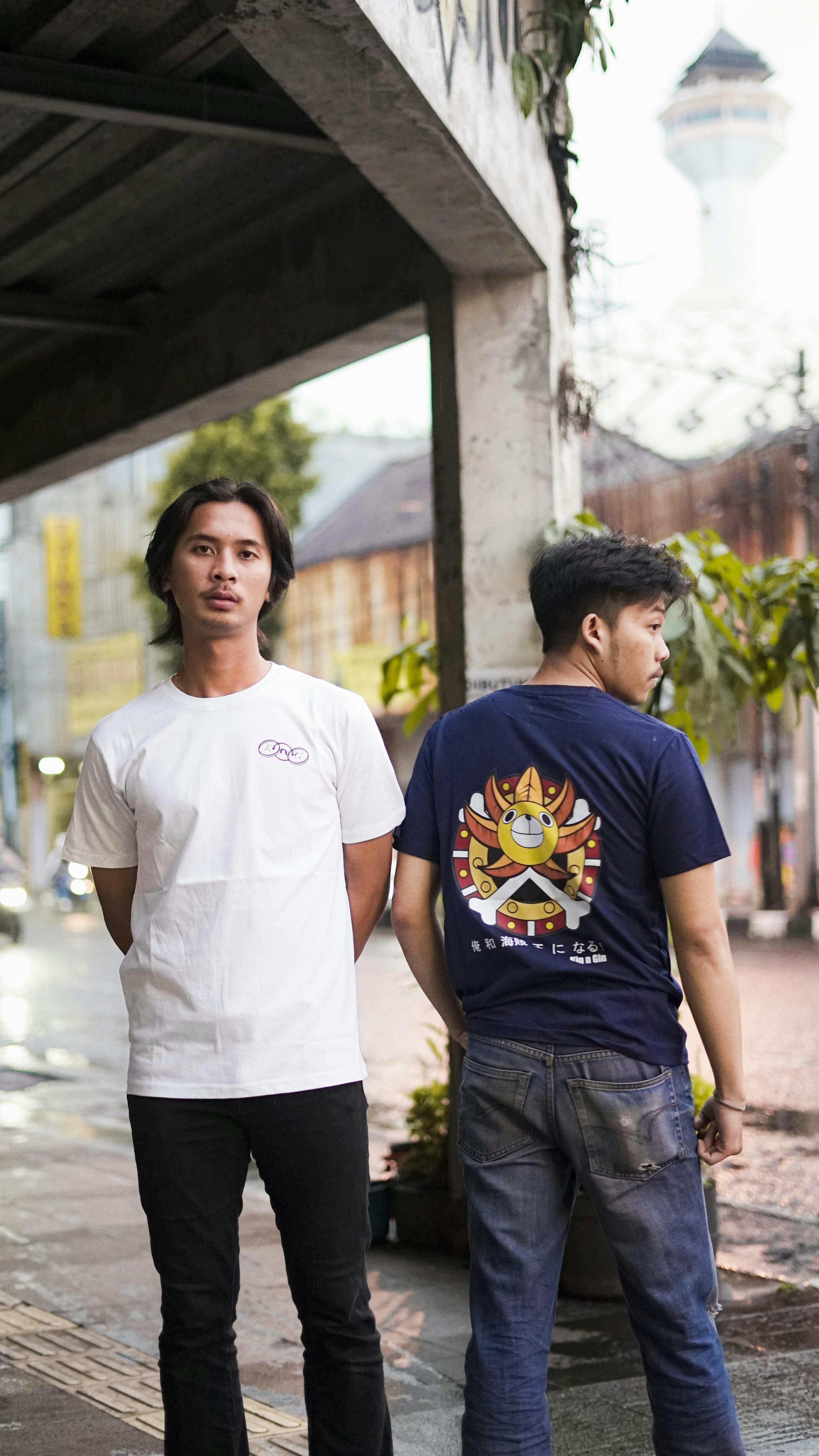 Two young Asian men in casual T-shirts standing on a city street under an overpass.