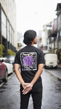 Back view of a young man wearing a trendy black T-shirt with a purple design on a city street.