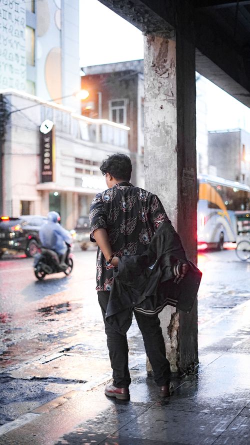 Free A man stands on a rainy city street holding a jacket, observing urban life. Stock Photo
