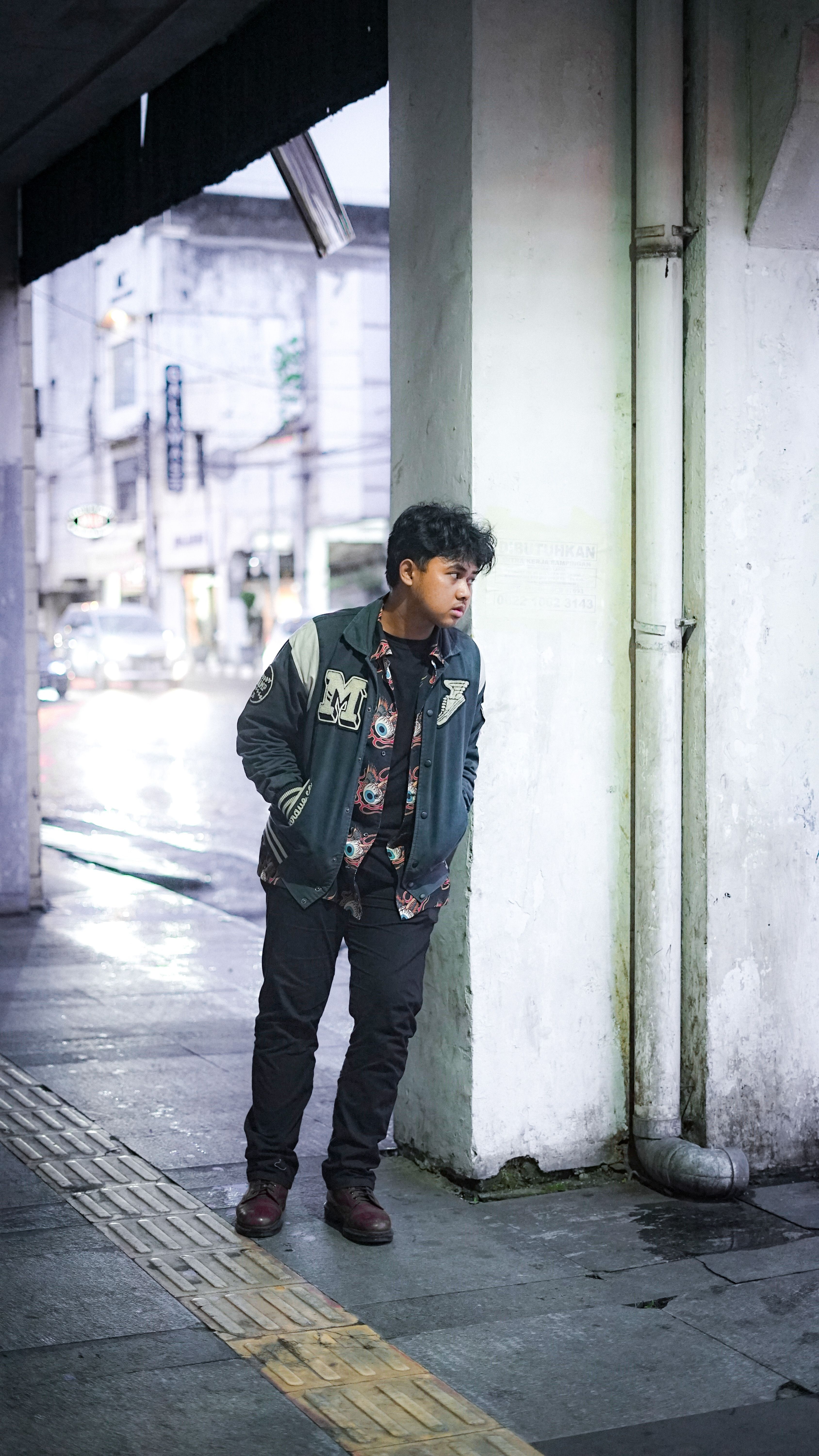 Boy leaning against a wall in a rain-soaked urban environment, wearing a casual jacket.