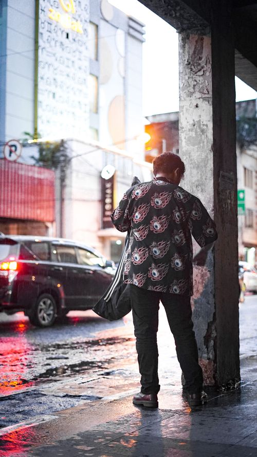 Free Back view of a man standing on a rainy city sidewalk with cars and an urban backdrop. Stock Photo