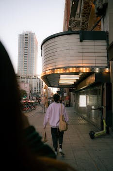 A pedestrian walks past a lit-up theater marquee on a city street during twilight.