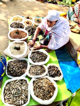 Woman vendor at outdoor market with an assortment of dried goods displayed for sale.