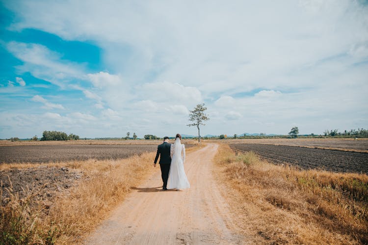 A Back View Of A Couple Walking On The Dirt Road Between Grass Field