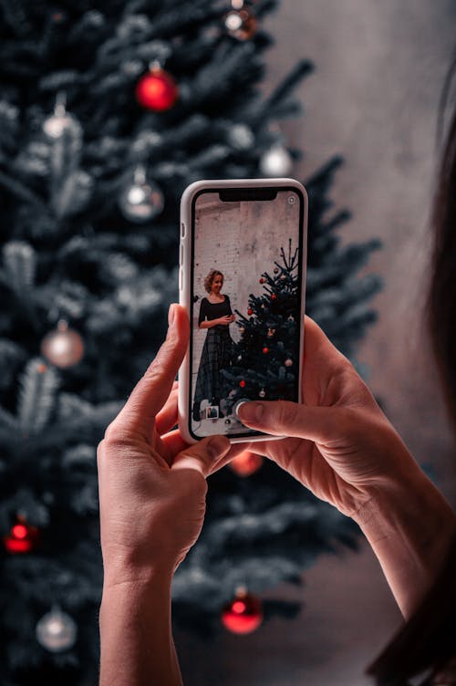 Person Taking Photo of Woman Standing Beside Christmas Tree