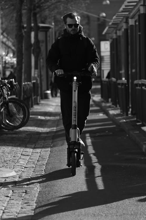 A Grayscale Photo of a Man Riding a Scooter on the Street