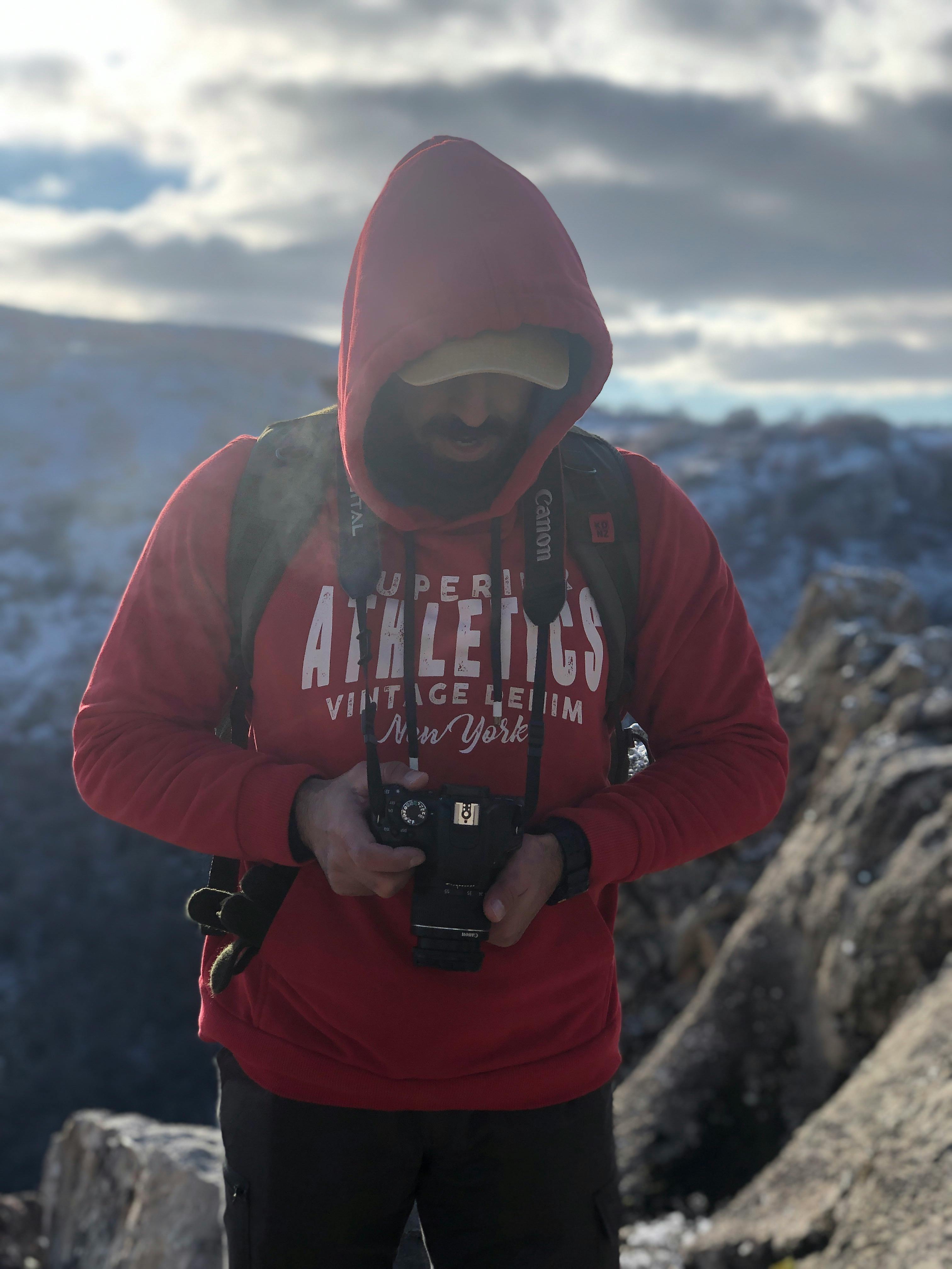 Man in red hoodie with camera on a mountain, capturing scenic views.