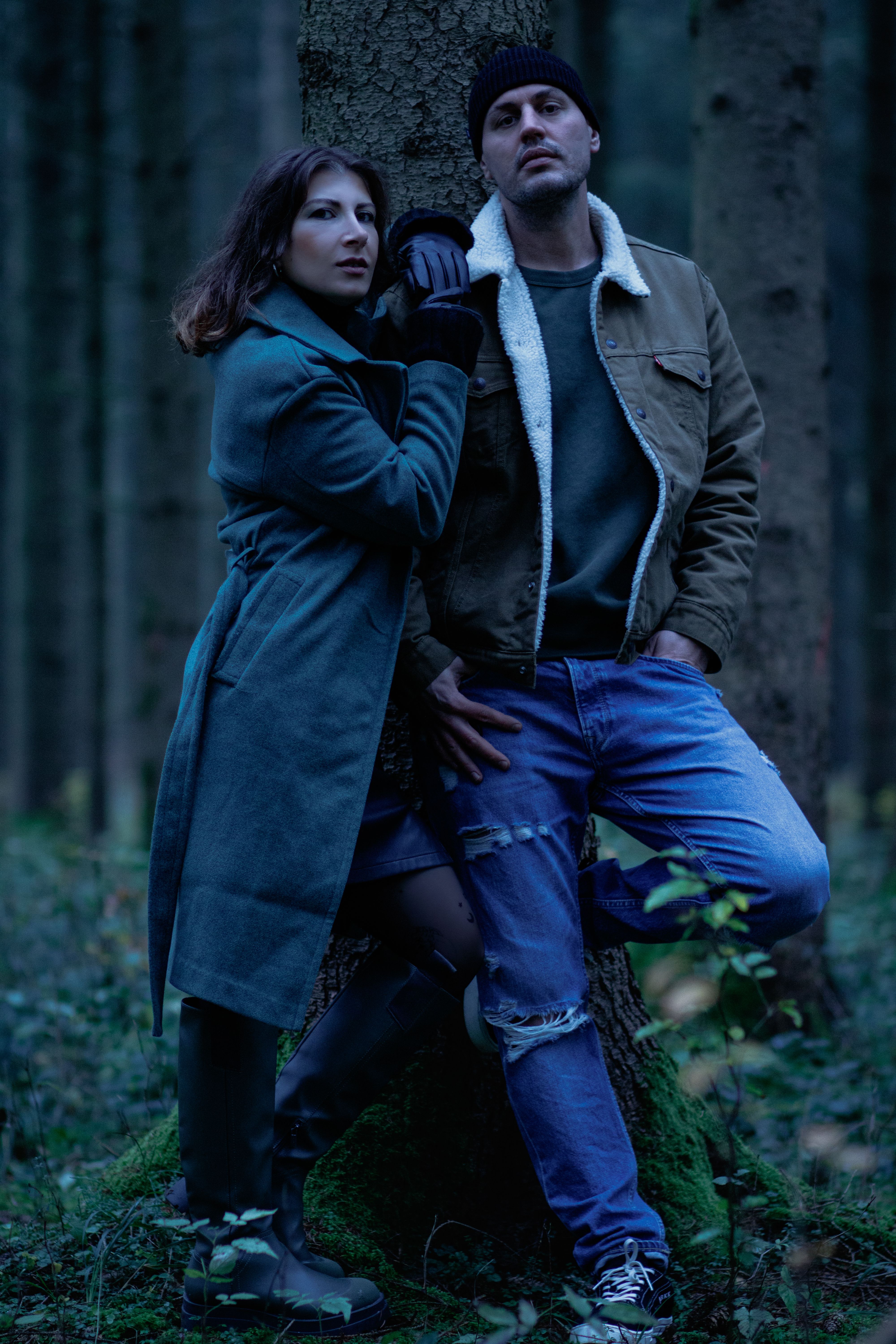 Stylish couple in coats posing in a serene forest, conveying a cool, modern vibe.