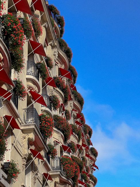 Luxury Parisian hotel exterior with red awnings and floral balconies against clear blue sky.