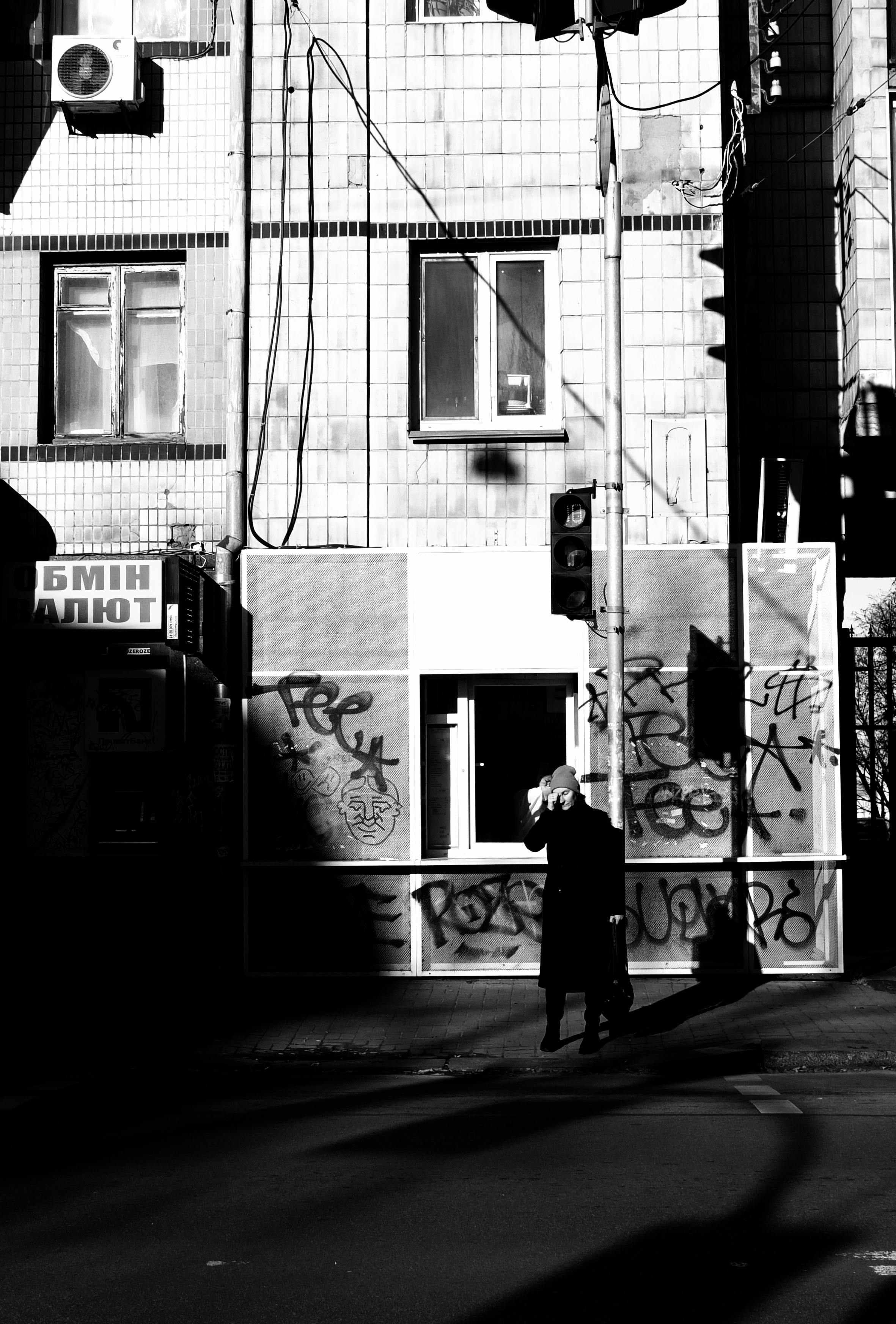 Black and white urban street photo showing a person walking in shadowed graffiti-covered area.