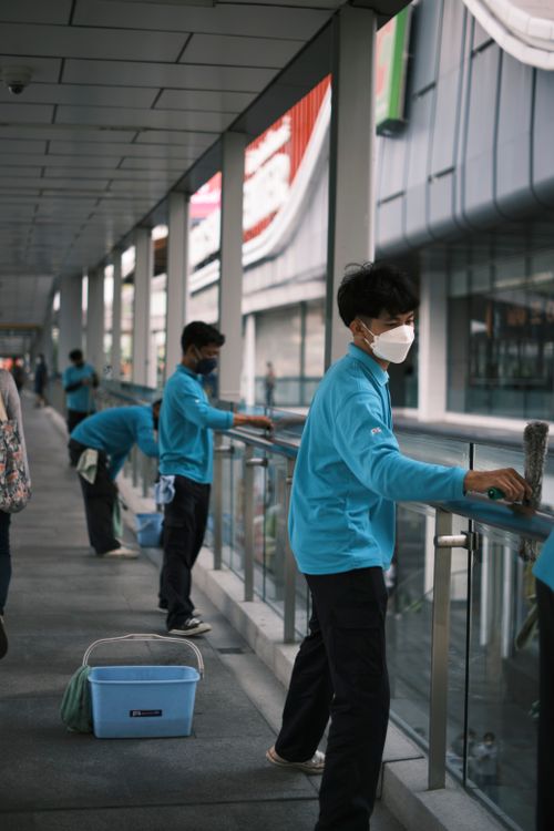 Free A team of workers in blue shirts with face masks clean glass railings outdoors during the day. Stock Photo