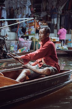Elderly man rowing a boat in a bustling Thai floating market in Bangkok.