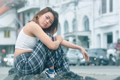 A Woman in White Tank Top and Plaid Pants Sitting on the Street