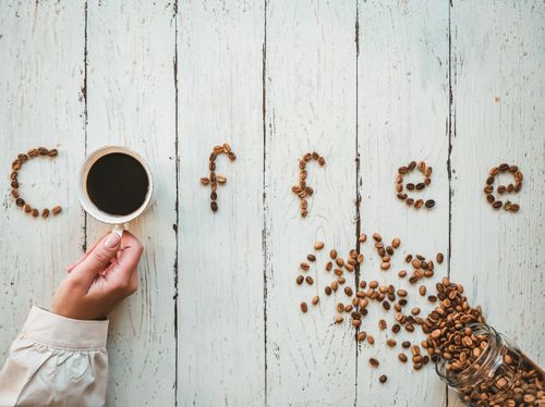 Free Creative coffee bean arrangement spelling 'coffee' on a rustic wooden table with a hand holding a cup. Stock Photo