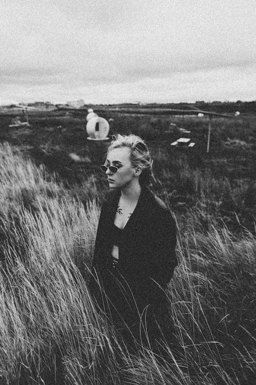 A Grayscale Photo of a Woman in Black Jacket Standing on Grass Field