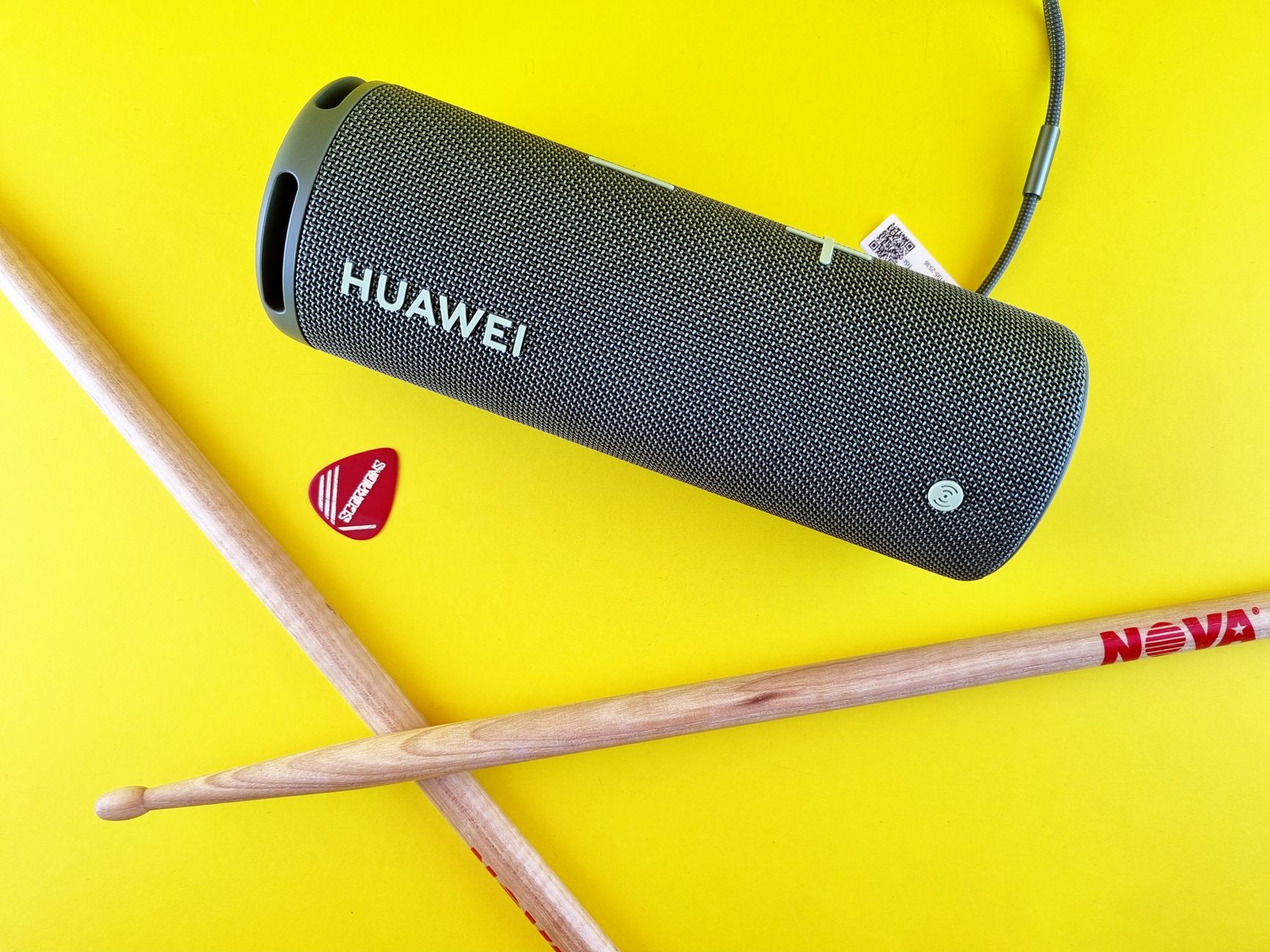 A Huawei speaker alongside drumsticks and guitar pick on a vibrant yellow surface.